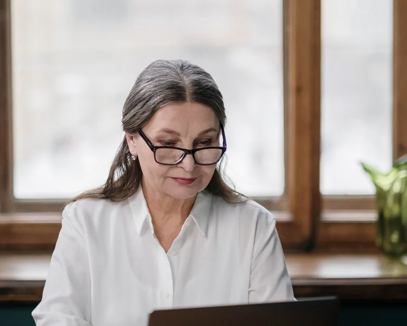 Idősebb nő szemüvegben, laptop előtt ül az asztalnál